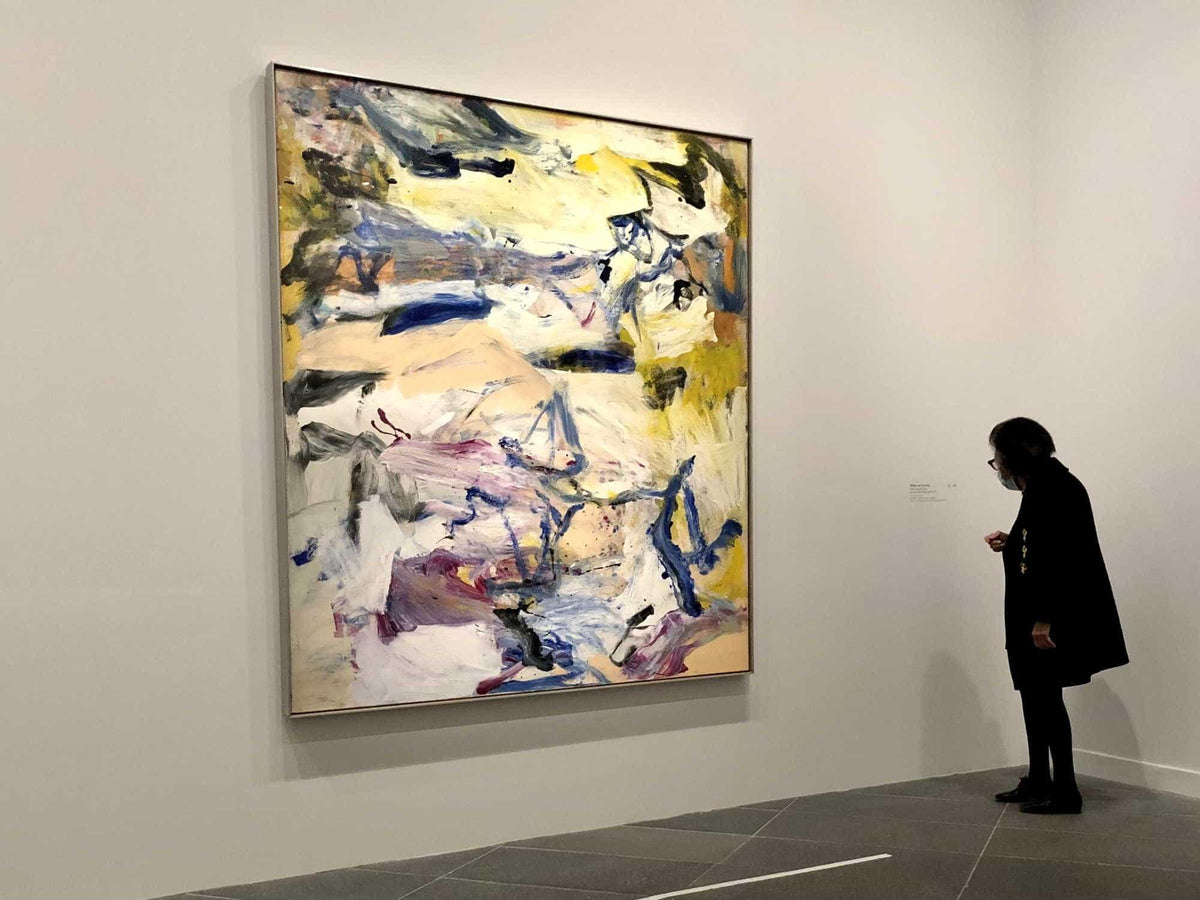 Woman viewing large abstract painting with bold brushstrokes in yellow, blue, and purple at art gallery