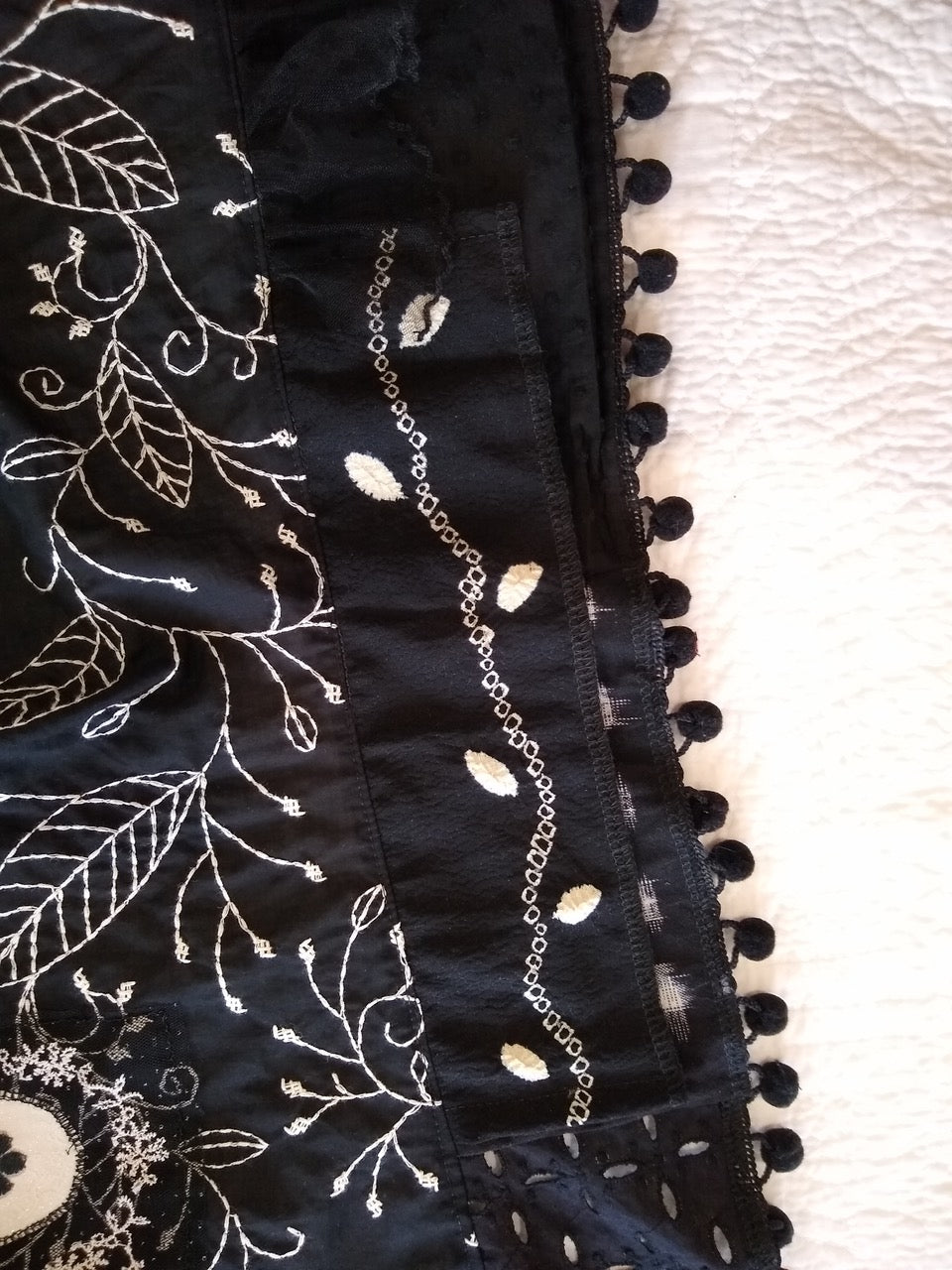 Black fabric with white leaf embroidery and pom-pom trim, ideal for apparel sewing projects.