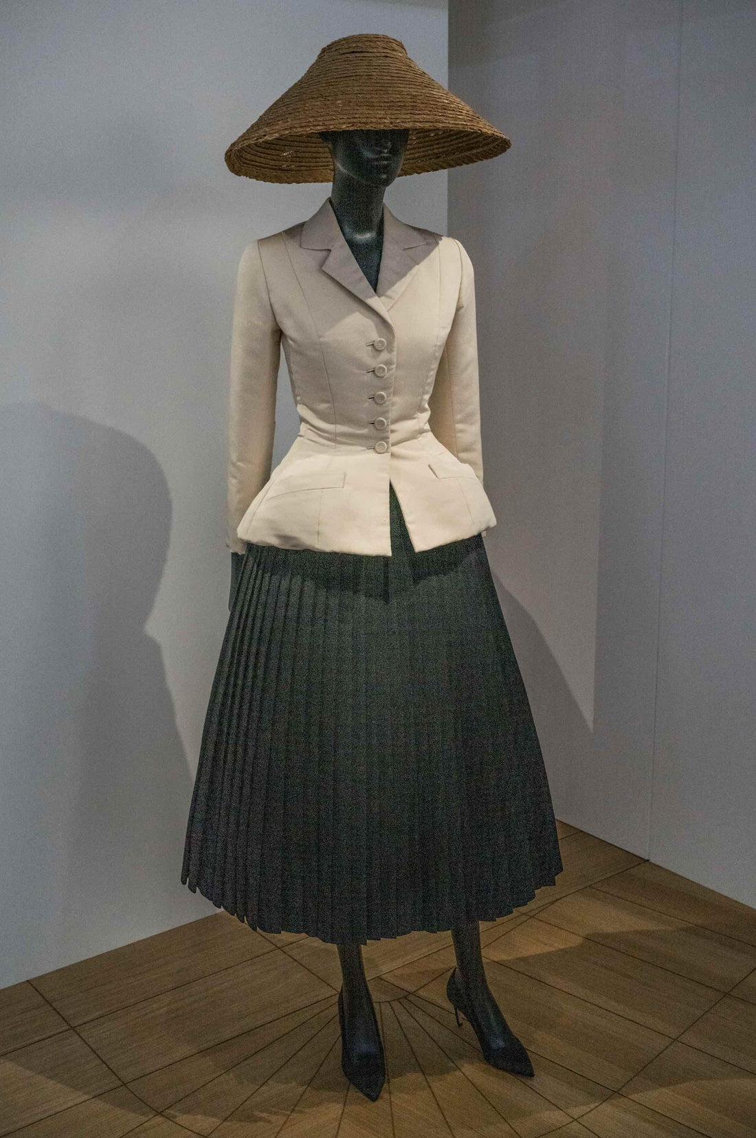 Cream tailored jacket with buttons, dark pleated skirt, and wide-brim straw hat on mannequin