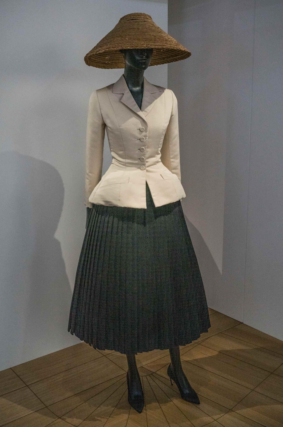 Cream tailored jacket with buttons, dark pleated skirt, and wide-brim straw hat on mannequin