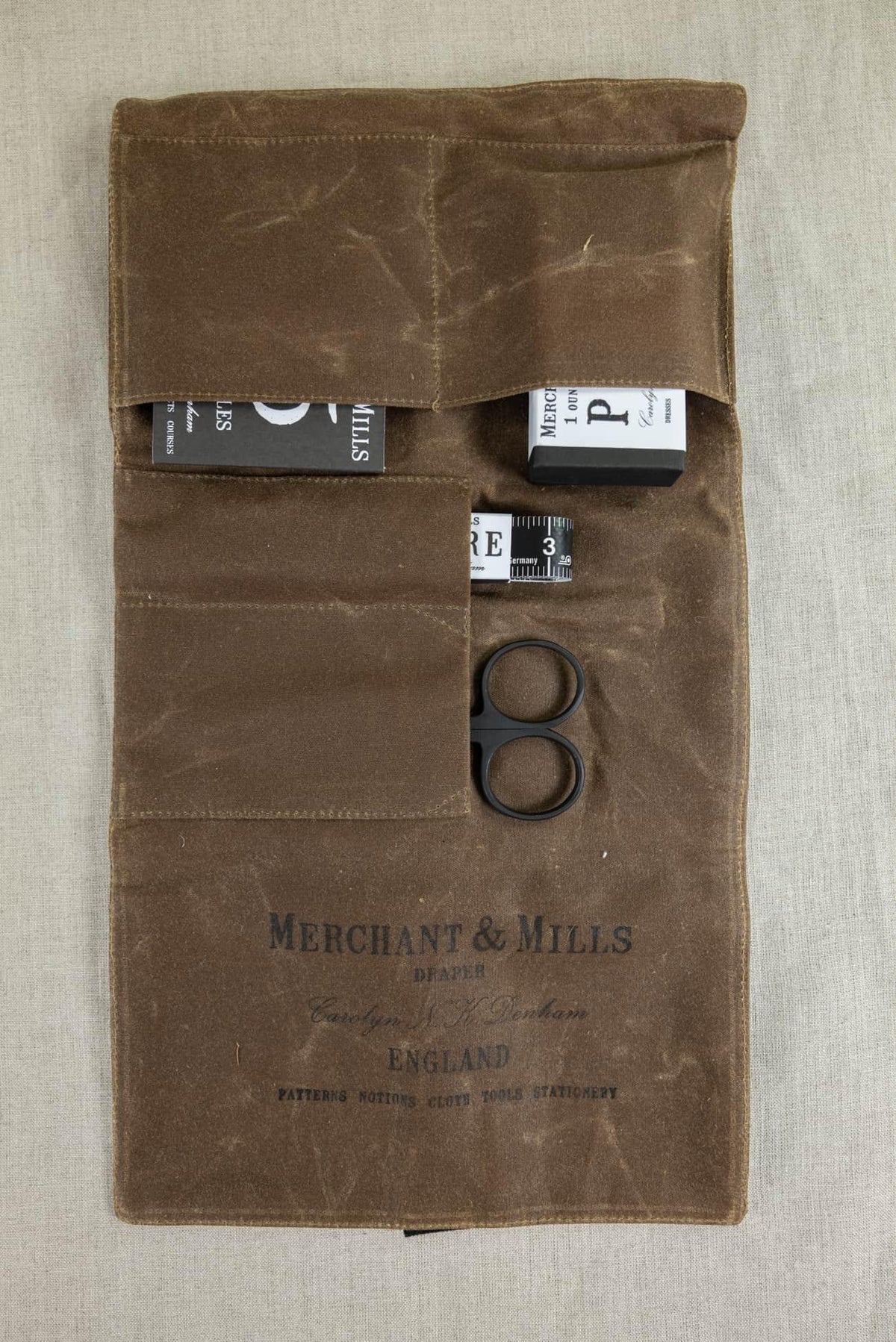 Brown waxed canvas sewing kit with Merchant & Mills tools, scissors, tape measure, and notions for creative sewing projects